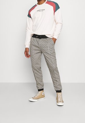 Crèmekleurig shirt met lange mouwen met teal- en bordeauxrode accenten op de schouders, gecombineerd met zwarte en witte joggers met patroon en beige hoge sneakers.