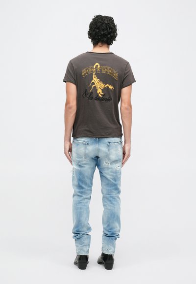 T-shirt cinza com impressão gráfica amarela nas costas, combinado com jeans claros desgastados e sapatos pretos, visto pela parte de trás.