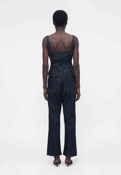 Γυναίκα που στέκεται με την πλάτη γυρισμένη, φοράει σκούρο αμάνικο τοπ από τζιν με δέσιμο στο πίσω μέρος και τζιν με ίσια μπατζάκια που ταιριάζει, καθώς και μαύρα σανδάλια με τακούνι.