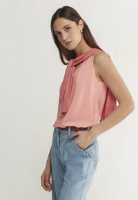 Ljusrosa ärmlös topp med rundad halsringning, tillsammans med vida ljusa denimbyxor. En mjuk rosa tröja hänger över axlarna.