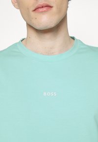 Ljus mintgrön bomullströja med rund halsringning och en liten vit "BOSS"-logotyp tryckt på bröstet. Slät textur, avslappnad design.