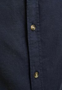 Camisa de lino azul marino con una tapeta de botones con botones dorados, tela texturizada y un cuello bien cuidado. Costuras visibles a lo largo de los bordes.