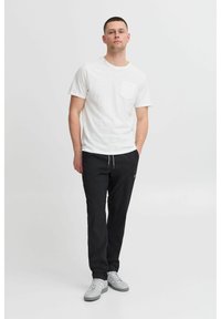 Camiseta blanca de manga corta con un bolsillo, combinada con pantalones negros con cordón y zapatillas grises. Diseño simple con un corte relajado.