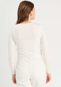 Light beige fitted long-sleeve top with a round neck, featuring a smooth texture. Worn with white high-waisted jeans and a subtle brand tag.