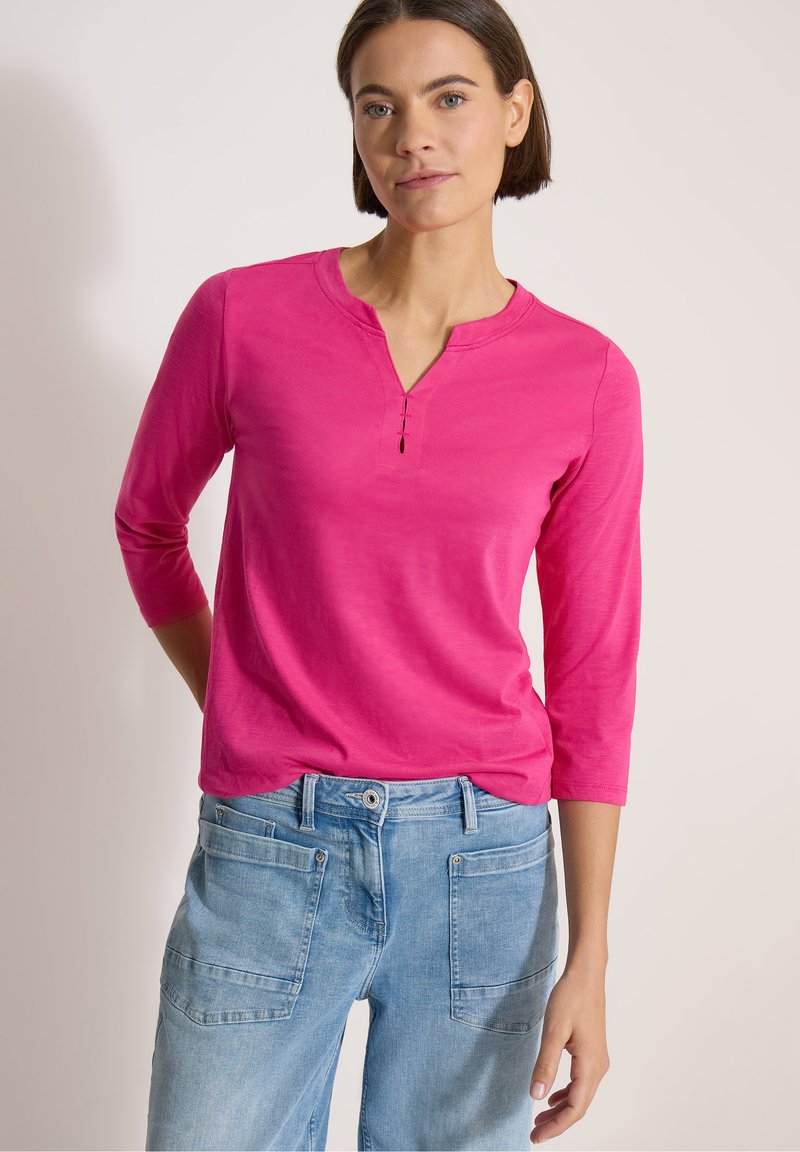 Femme aux cheveux courts et bruns portant un haut rose vif à manches trois-quarts et un jean en denim bleu clair sur un fond uni.