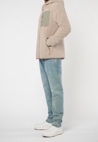 Personne portant une veste en polaire beige avec une poche poitrine verte, un jean bleu clair et des baskets blanches, debout devant un fond clair uni.