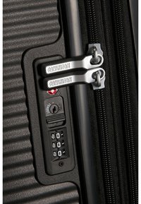 Valigia rigida nera con texture a coste, doppie cerniere argentate, lucchetto TSA e dials combinatori. Caratteristiche di design compatto e durevole.