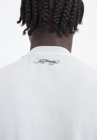 Světle šedé tričko s krátkým rukávem a kulatým výstřihem, s černým vyšitým logem "Ed Hardy" na zátylku.