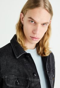 Black denim jacket with button closures and two chest pockets, worn over a light blue shirt. Features a standard collar and textured fabric.