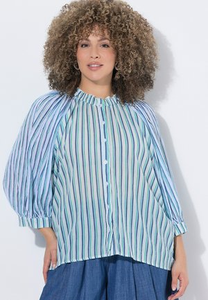 Kvinde med krøllet hår iført en knaplukket, langærmet, stribet bluse med puffede ærmer og løse blå bukser, stående mod en hvid væg.