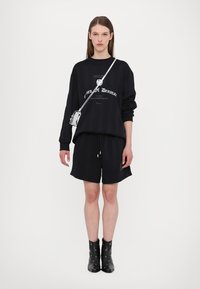 Black oversized sweatshirt with front graphic, paired with matching black shorts. Model wears black ankle boots with eyelet accents and a silver crossbody bag.