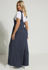 Mörk denim jumpsuit med vida ben, ribbad textur och justerbara axelband. Matcher med en vit t-shirt och vita sneakers.