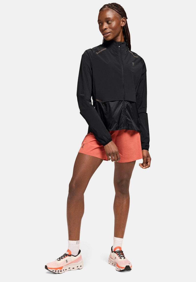 Chaqueta de running negra con cremallera en la parte delantera, mangas largas y un panel inferior de malla, combinada con pantalones cortos coral y zapatillas rosas.