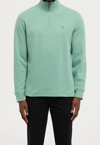 Man i mintgrön pullover med kvartdragkedja och en liten broderad logotyp på bröstet, i svarta byxor, st ående med avslappnade armar.