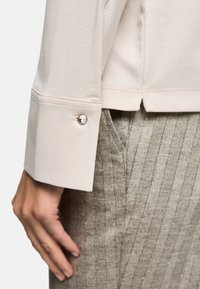 Light beige long-sleeve top with a cuff featuring a decorative silver button. The fabric is smooth, paired with textured gray, pinstriped pants.