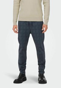 Graue karierte Hose aus weichem Material, mit einem elastischen Bund und dezenten Falten; kombiniert mit schwarzen Stiefeletten und einem beigen Oberteil.