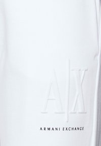Vita bomullsjoggers med en präglad logotyp "A|X" och texten "ARMANI EXCHANGE" i svart. Slät textur med en modern design.
