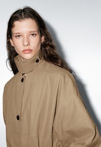 Beige, lichtgewicht jas met een hoge kraag, grote knopen en een relaxte pasvorm. Gladde textuur; heeft een subtiele glans en strakke lijnen.