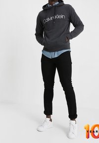 Felpa grigia con cappuccio e logo "Calvin Klein" bianco, indossata sopra una camicia chambray azzurro chiaro, pantaloni neri a sigaretta e sneakers bianche.