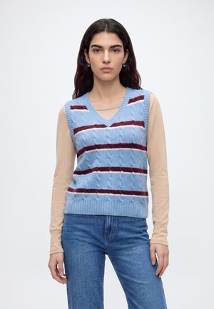 Jeune femme aux longs cheveux foncés portant un pull sans manches bleu torsadé avec des rayures bordeaux sur un top beige à manches longues et un jean bleu.