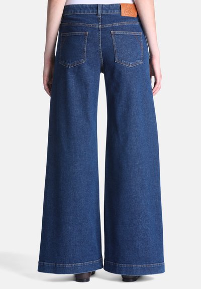 Personne portant un jean bleu à jambes larges avec poches arrière et un patch en cuir de marque, debout devant un fond blanc uni.