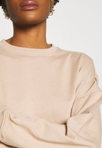 Sweatshirt beige clair avec un col rond, des poignets côtelés et une texture douce. Associé à des boucles d'oreilles créoles en métal doré, ajoutant de l'éclat.