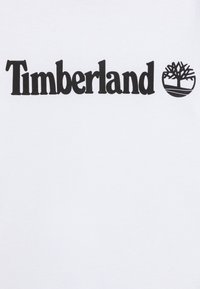 Camiseta blanca de algodón con el logo de "Timberland" en una fuente negra en negrita, acompañado de un diseño estilizado de un árbol y un gráfico de base circular.