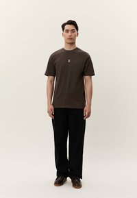 Ung man som står rakt upp, klädd i en mörkbrun t-shirt med "II"-logotyp, svarta byxor och bruna skor mot en enkel ljus bakgrund.