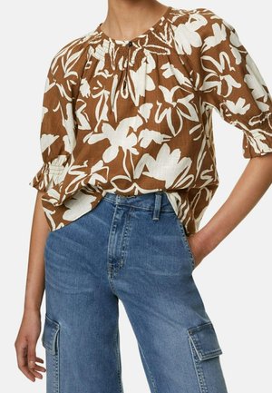 Personne portant un chemisier à manches bouffantes à fleurs marron et blanc rentré dans un jean cargo en denim bleu, les mains dans les poches.