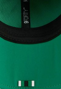 Grüne 9FORTY-Kappe mit einem strukturierten Mesh-Innenfutter und einem schwarzen Schirm. Sie verfügt über ein dreifärbiges Akzentdetail am unteren Rand: grün, schwarz und weiß.