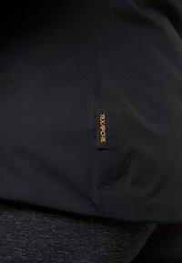 Svart textiljacka med texturerad yta, med en gul "TEXAPORE"-etikett på nederkanten. Designen är smidig och funktionell.