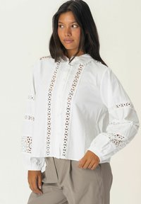 White blouse featuring intricate lace cutouts along the front and sleeves, with a collared neck and button closure. Slightly loose fit.