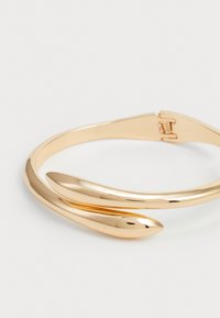 Gold-tone metal bangle with a two-curved design. Smooth, shiny surface with tapered ends, featuring a secure clasp closure.
