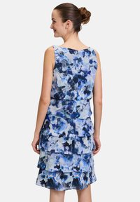 Sukienka w kwiatowy wzór w odcieniach niebieskiego, bez rękawów, z warstwowymi falbanami. Materiał wydaje się lekki i zwiewny, z zauważalnymi różnicami w fakturze i wzorze.