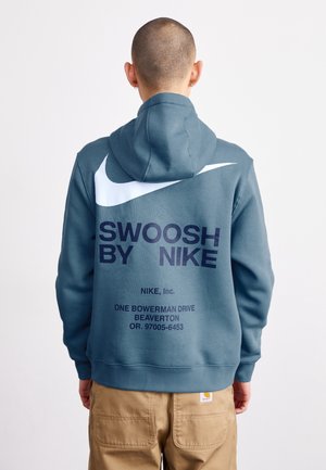 Osoba z ogoloną głową ubrana w niebieską bluzę z kapturem Nike z białym znakiem i napisem "SWOOSH BY NIKE" na plecach oraz beżowe spodnie.