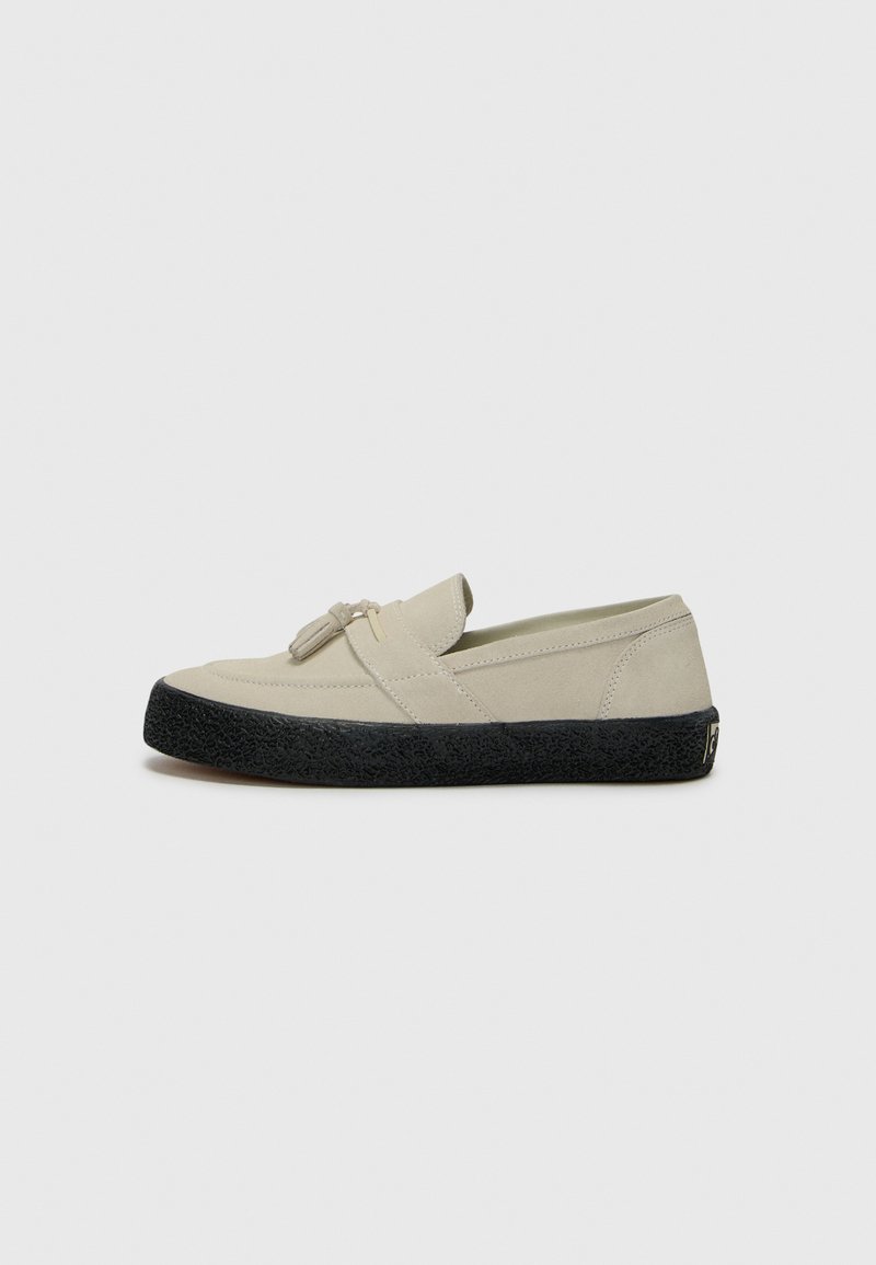 Light beige suede slip-on shoe with rounded toe, black textured rubber sole, and decorative tassel detail on the front.