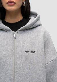Felpa grigia con cerniera, caratterizzata da un ampio cappuccio, una texture morbida e il logo ricamato "WRSTBHRV" sul lato sinistro. Design semplice e vestibilità casual.