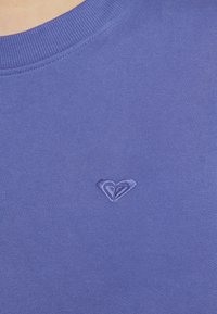 Primo piano di una persona che indossa una maglietta a girocollo a coste di colore viola con un piccolo logo a forma di cuore ricamato sul petto.