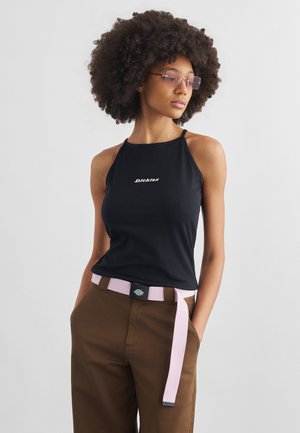 Γυναίκα με αφρο χτένισμα, που φοράει μαύρη αμάνικη μπλούζα, καφέ παντελόνι, ροζ ζώνη και φιμέ γυαλιά, στέκεται με τα χέρια στις τσέπες.