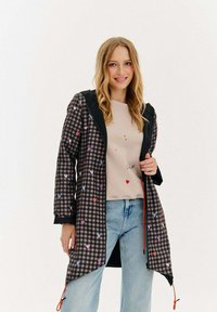 Sieviete smaida, ģērbta melnā šaha raksta kapucē ar daudzkrāsainām sirdīm, bēšā topā un gaiši zilos džinsos uz vienkārša balta fona.