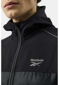 Czarna kurtka Reebok z wysokim kołnierzem, zapinana na zamek, o kontrastowej, błyszczącej strukturze materiału. Posiada srebrne logo na klatce piersiowej oraz odblaskowe akcenty.