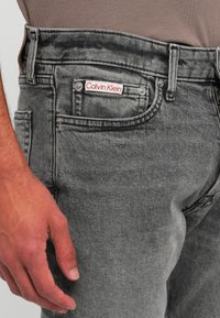 Man’s hand resting beside faded gray denim jeans with visible Calvin Klein logo tag on the front pocket.
