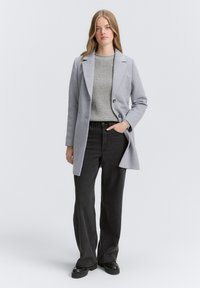 Grijze wolletjesjas met een enkelvoudige sluiting, met klepjes en twee knopen. Gecombineerd met een grijze geribbelde trui en zwarte wijd-uitlopende broek.
