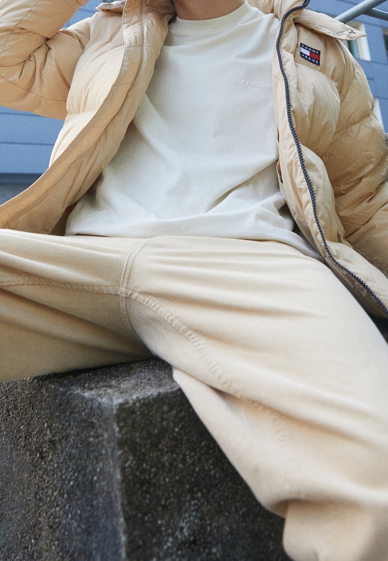 Veste matelassée beige, t-shirt crème clair et pantalon assorti. La veste présente un logo et un tissu doux texturé avec des coutures visibles.