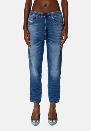 Przedni widok osoby noszącej wyblakłe niebieskie dżinsowe spodnie dresowe z elastycznym pasem i sznurkiem, w połączeniu z błyszczącymi butami na obcasie o szpiczastym nosku.