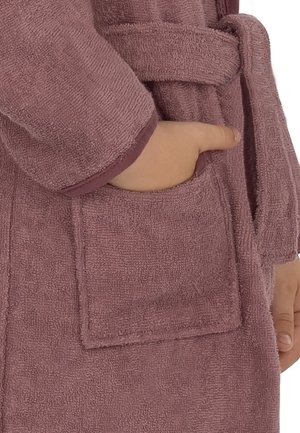 Kinderhand, die in die Tasche eines strukturierten, mauvefarbenen Bademantels steckt, der mit einem passenden Gürtel in der Taille gebunden ist.