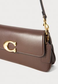 Sac à épaule en cuir marron avec un fermoir doré "C" et une quincaillerie assortie, avec une étiquette en cuir attachée par une chaîne dorée.