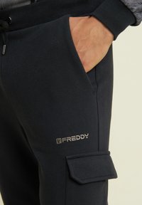 Sudadera de color azul marino oscuro con un bolsillo con cremallera y un logo en el muslo. Está hecha de una tela suave con una textura lisa y puños elásticos.