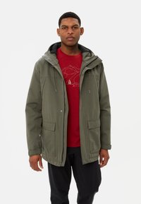 Giacca verde oliva con cappuccio, dotata di chiusura con zip e bottoni a pressione, due tasche frontali e un morbido interno foderato in nero. Camicia rossa sotto.
