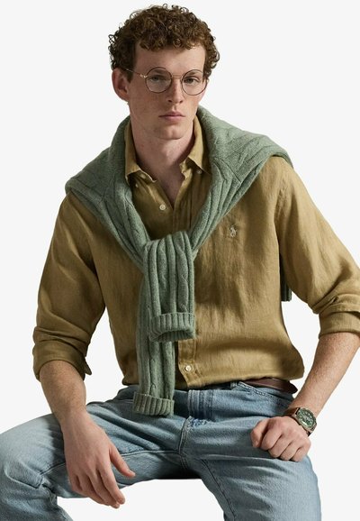 Jeune homme aux cheveux bouclés portant des lunettes rondes, une chemise moutarde, un jean bleu clair, un pull vert drapé sur les épaules et une montre-bracelet.